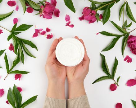 two female hands and a jar with thick cream and burgundy flowering peonies with green leaves, top view, concept of anti-aging procedures for rejuvenating and moisturizing the skin of hands