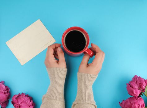 red creameric cup with black coffee in female hands and a bouquet of red peonies with green leaves on a blue background, top view