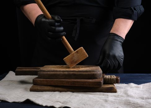 male cook in black uniform holds a wooden hammer for beating meat on a vintage cutting board, black background