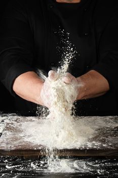 chef in black uniform sprinkles white wheat flour in different directions, product scatters dust, black background