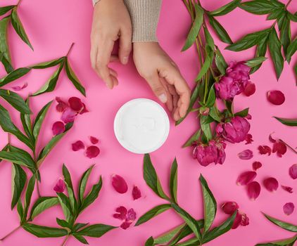 two hands of a young girl with smooth skin and a jar with thick cream and burgundy  peonies with green leaves, top view, concept of anti-aging procedures for rejuvenating and moisturizing 