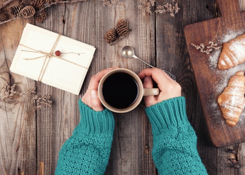 women's hands in a green knitted sweater holding a ceramic mug with black coffee, next to baked croissants sprinkled with powdered sugar, gray wooden background, top view