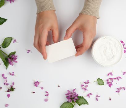 female hands and a jar with thick cream and pink flowers with green leaves, top view, concept of anti-aging procedures for rejuvenating and moisturizing the skin 