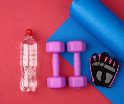 bottle of water, blue twisted neoprene mat, pair dumbbells and gloves on a red texture background, top view, sports set