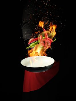 chef in a black uniform holds a round pan and throws up red and green whole chili peppers in a burning fire, cooking spices, low key