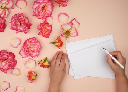 female hand holds a white pen over empty white sheet of paper, next to a bouquet of blooming pink roses, top view