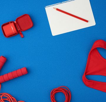red sports shirt, red jump rope and wireless headphones in a red case on a blue background, top view, empty white sheet of paper, flat lay