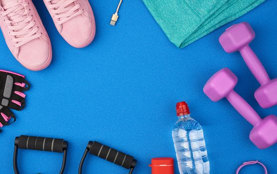 plastic purple dumbbells, sportswear, water, pink sneakers and wireless headphones on a blue background from a sports mat, fitness kit, flat lay, copy space