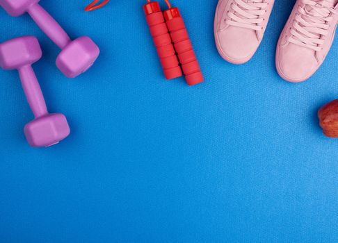 pair of pink suede sneakers, dumbbells and a red jump rope on a blue background, top view, set for sports, copy space