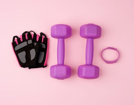 pair of purple plastic dumbbells and black gloves on a pink background, training equipment, top view