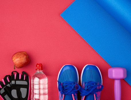 pair of blue training sneakers with laces, bottle of water and a blue twisted neoprene mat on a red texture background, top view, sports set