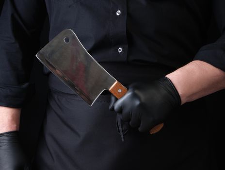male cook in black uniform and black latex gloves holds a large sharp meat knife, close up