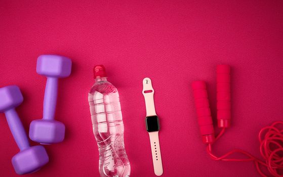 smart watch, bottle of water, red jump rope and lilac dumbbells on a red background from a mat for yoga, top view, sports backdrop