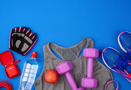 plastic purple dumbbells, sportswear, water, pink sneakers and wireless headphones on a blue background from a sports mat, fitness kit, flat lay, copy space