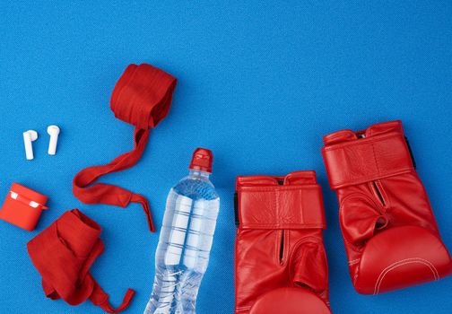 pair of red leather boxing gloves, a textile red elastic bandage for hands, wireless headphones and a bottle of water lie on a neoprene blue mat, sports kit