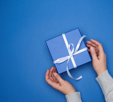 two female hands in a sweater hold a square blue box with a bow on a blue classic background, top view, festive backdrop