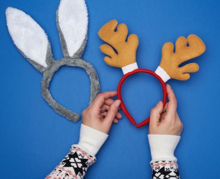 female hands holding carnival masks of fur, hoop with rabbit and horned deer ears on a blue background, top view