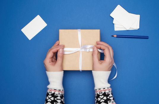 two female hands are holding a cardboard gift square box tied with a white ribbon and empty paper business cards on a dark blue background, top view, classic trend colors