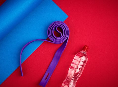 pair of plastic dumbbells, purple elastic rubber and bottle of water on a red neoprene mat, top view, training equipment