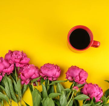 red creameric cup with black coffee and a bouquet of red peonies with green leaves on a yellow background, top view