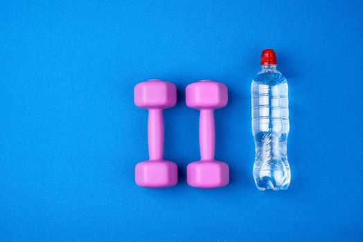 transparent plastic bottle of water and a pair of purple plastic dumbbells for sports and fitness on a blue mat, top view, copy space