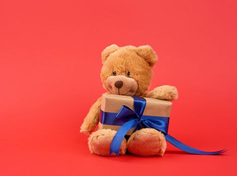 cute little brown teddy bear holds a brown box with a blue ribbon on a red background, valentine's day card