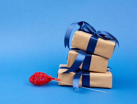 stack of gifts wrapped in brown kraft paper and a silk blue ribbon, next to a red decorative heart, blue background