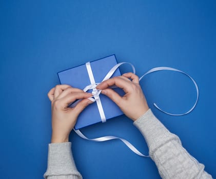 two female hands in a sweater hold a square blue box with a bow on a blue classic background, top view, festive backdrop