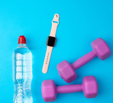purple plastic kettlebells, a transparent bottle of water and a smart watch on a blue background,  set for sports, top view, flat lay