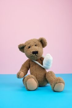 funny vintage brown curly teddy bear with rewound paw with white gauze bandage sitting on blue background, concept of injuries in children or animals