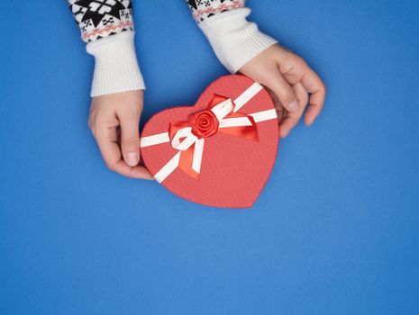 two female hands hold a paper closed red box in the shape of a heart on a dark blue background, festive backdrop, copy space