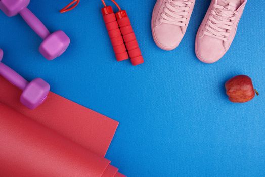 pair of pink training sneakers with laces, purple plastic dumbbells, red twisted neoprene mat on a blue texture background, top view, sports set