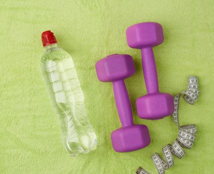 purple dumbbells and a clear plastic bottle of water on a green terry towel, top view, flat lay