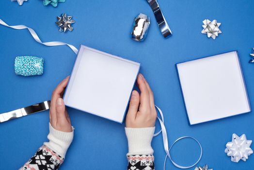 two female hands in a sweater hold a open square blue box with a bow on a blue classic background, top view, festive backdrop