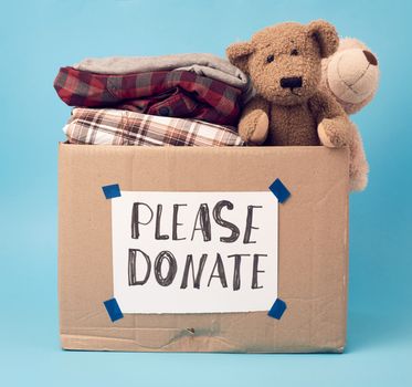 large brown cardboard box with the inscription please donate, box is filled with various clothes, toys on a blue background