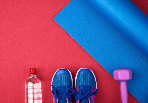 pair of blue training sneakers with laces, bottle of water and a blue twisted neoprene mat on a red texture background, top view, sports set