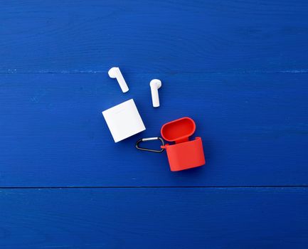 white wireless headphones, charging box and red silicone case on a blue wooden background, top view