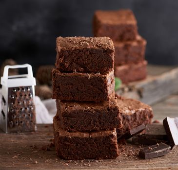 stack of baked square pieces of chocolate brownie cake on brown wooden cutting board, black background