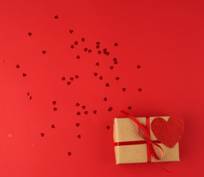 box wrapped in brown kraft paper and tied with a red thin silk ribbon, scattered confetti in the shape of hearts, red background, festive backdrop for Valentine's Day