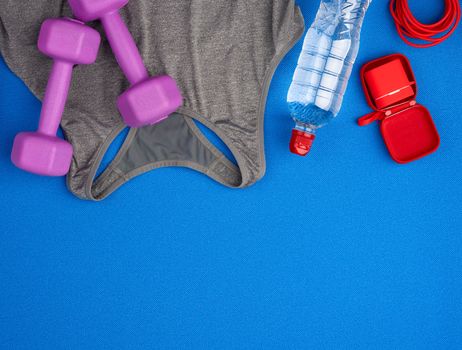 pair of purple plastic dumbbells, gray sports shirt and bottle of water on a blue background, set for sports, flat lay, copy space
