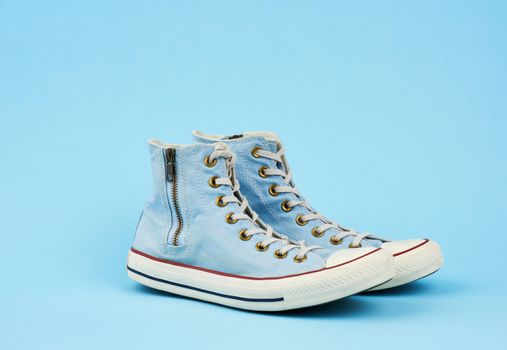 pair of light blue worn textile sneakers with laces and zippers on a blue background, side view of shoes