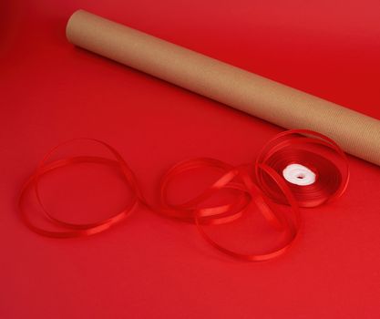 roll of brown paper for packaging, a reel of red ribbon and a wrapped gift in brown kraft paper on a red background, making surprises