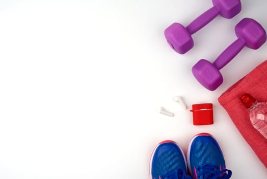 plastic purple dumbbells, bottle of water, blue sneakers and wireless headphones on a white background, fitness kit, flat lay, copy space