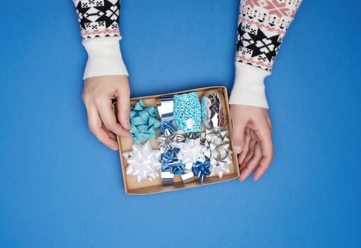 two female hands are holding a brown cardboard box with a set of decor ribbons and bows for wrapping gifts on a dark blue background, top view