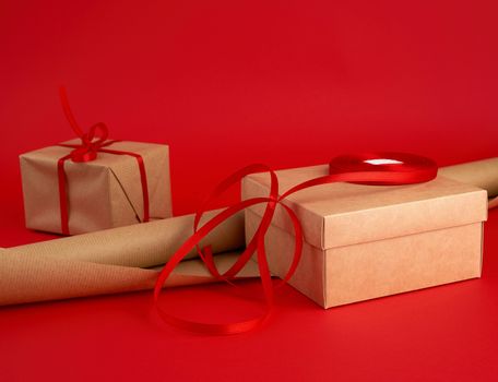 roll of brown paper for packaging, a reel of red ribbon and a wrapped gift in brown kraft paper on a red background, making surprises