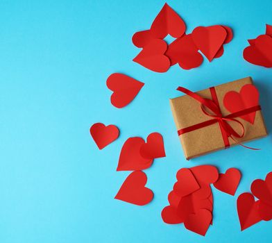rectangular box is wrapped in brown paper and tied with a red thin silk ribbon on a blue background, red cut out paper hearts, copy space, holiday is Valentine's Day