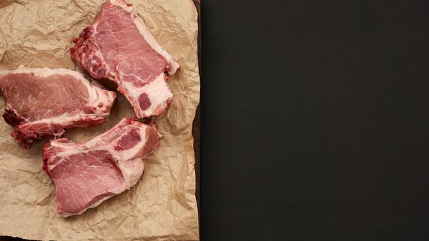 raw juicy pork slices of meat on the rib, food lies on brown parchment paper,  black wooden background and an empty space for writing a recipe or menu, top view