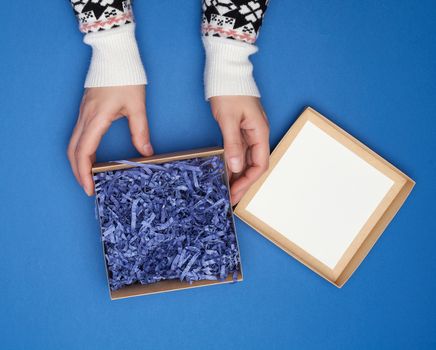 two female hands in a white sweater hold open square brown box on a blue classic background, top view