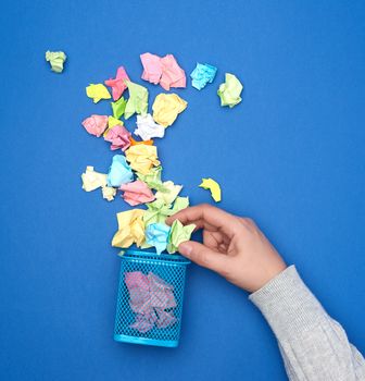 female hand holds crumpled multi-colored pieces of paper, next to a metal garbage bin, top view
