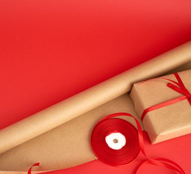 roll of brown paper for packaging, a reel of red ribbon and a wrapped gift in brown kraft paper on a red background, making surprises, place for an inscription